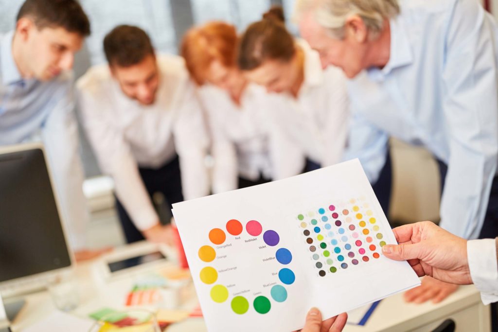 Mehrere Menschen in Bürokleidung beugen sich über Tisch, auf dem Farbsamples liegen